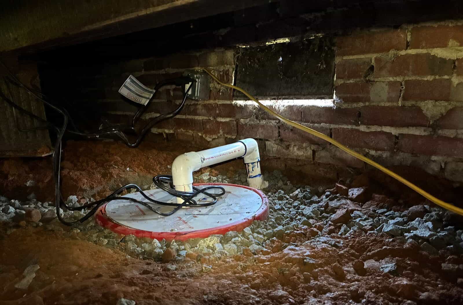 Sump basin with a sealed lid and white discharge pipe set in gravel in a crawl space.