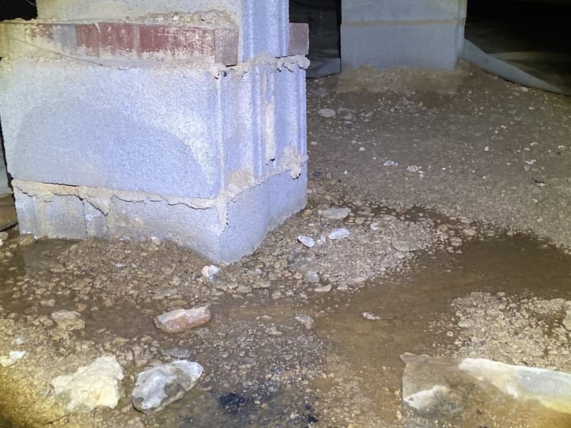 Standing water on the dirt floor of a crawl space beside a block pier