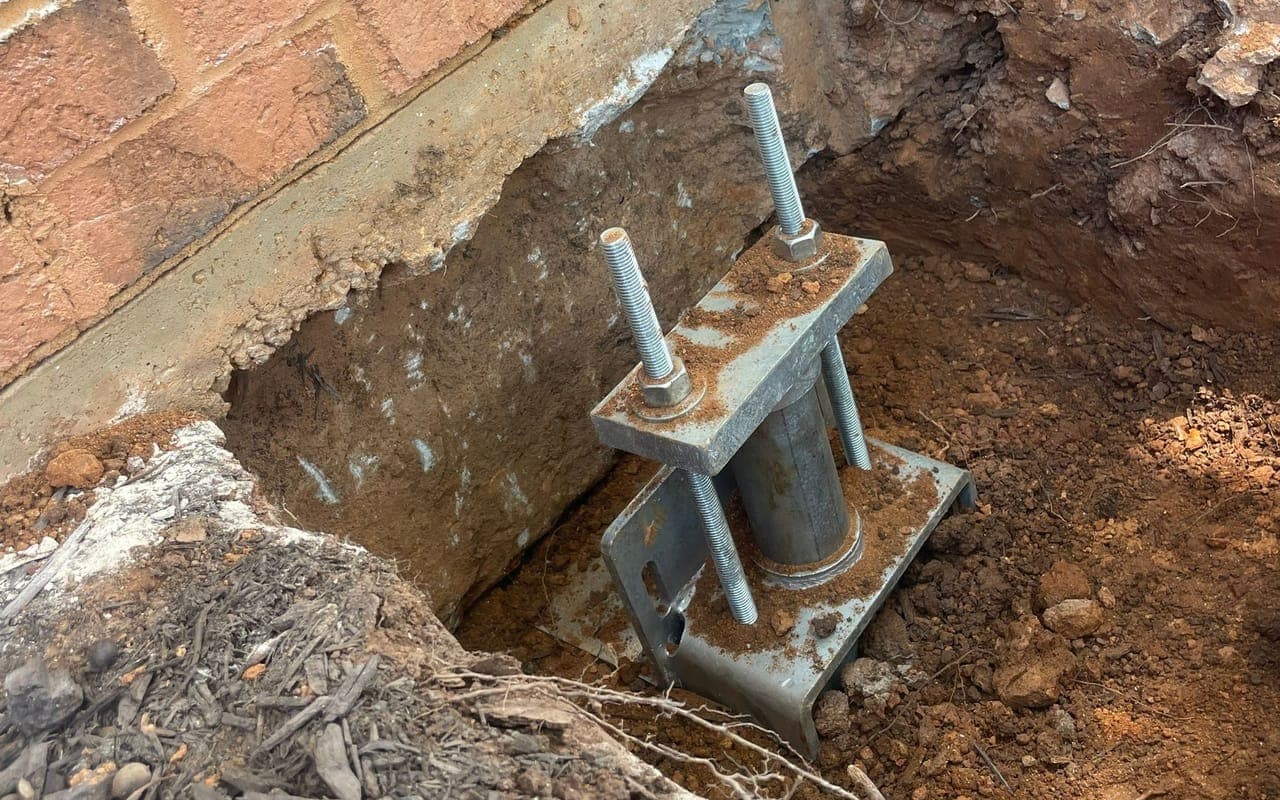 A helical pier bracket set at the footing of a brick home in an open excavation