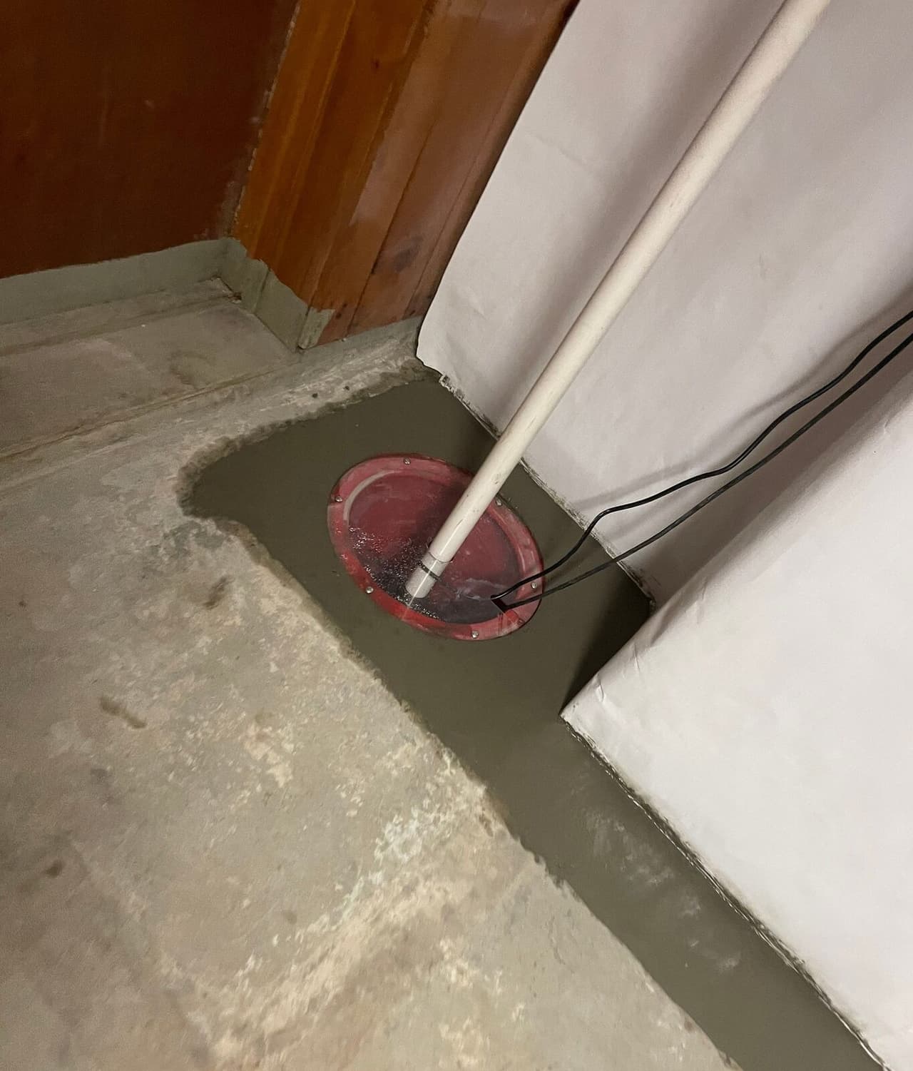 Sump basin with red lid and discharge pipe set in fresh concrete patch beside white wall liner.