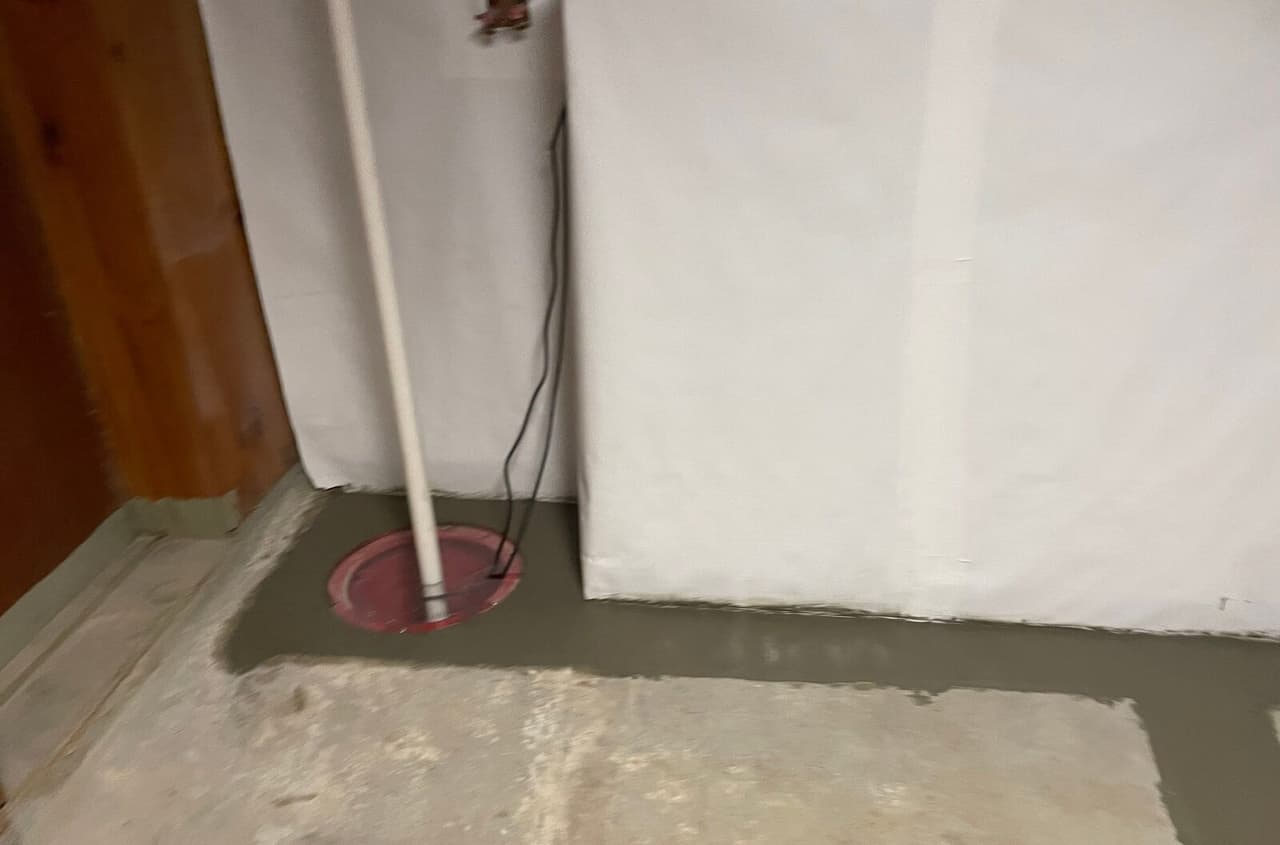Sump basin with red lid and pipe set in fresh concrete at the base of white lined basement walls.