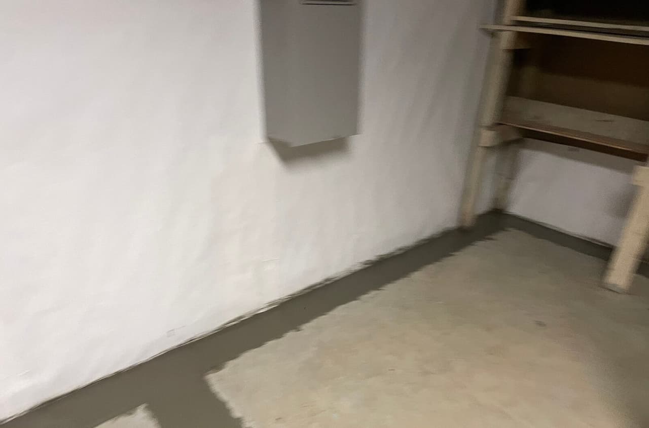 Blurry view of a white lined basement wall with a fresh concrete strip over the drain along the floor.