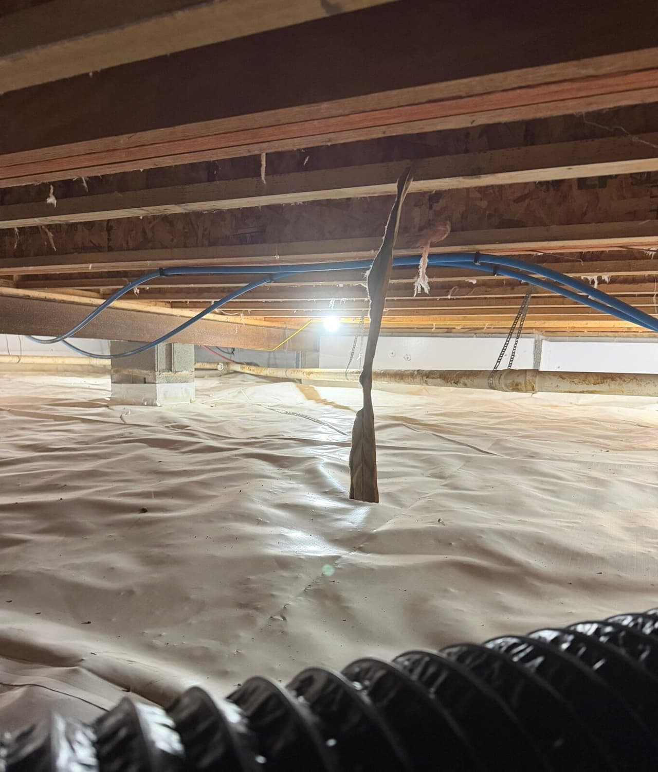 Crawl space with white liner, wrapped block pier, blue lines overhead and a black duct in front.