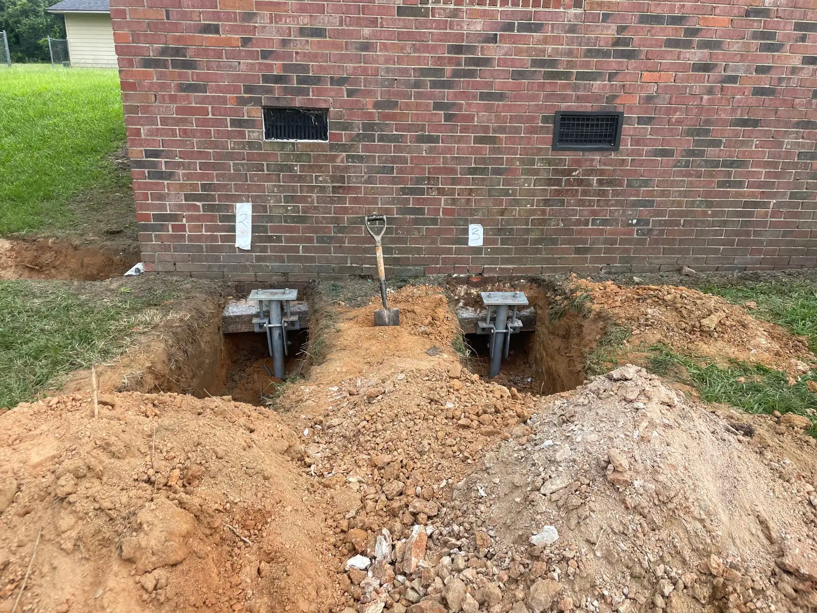 Two steel pier brackets in excavated holes along the foundation of a brick home