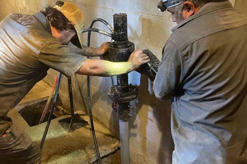 Two crew members driving a steel pier shaft into the soil inside a basement
