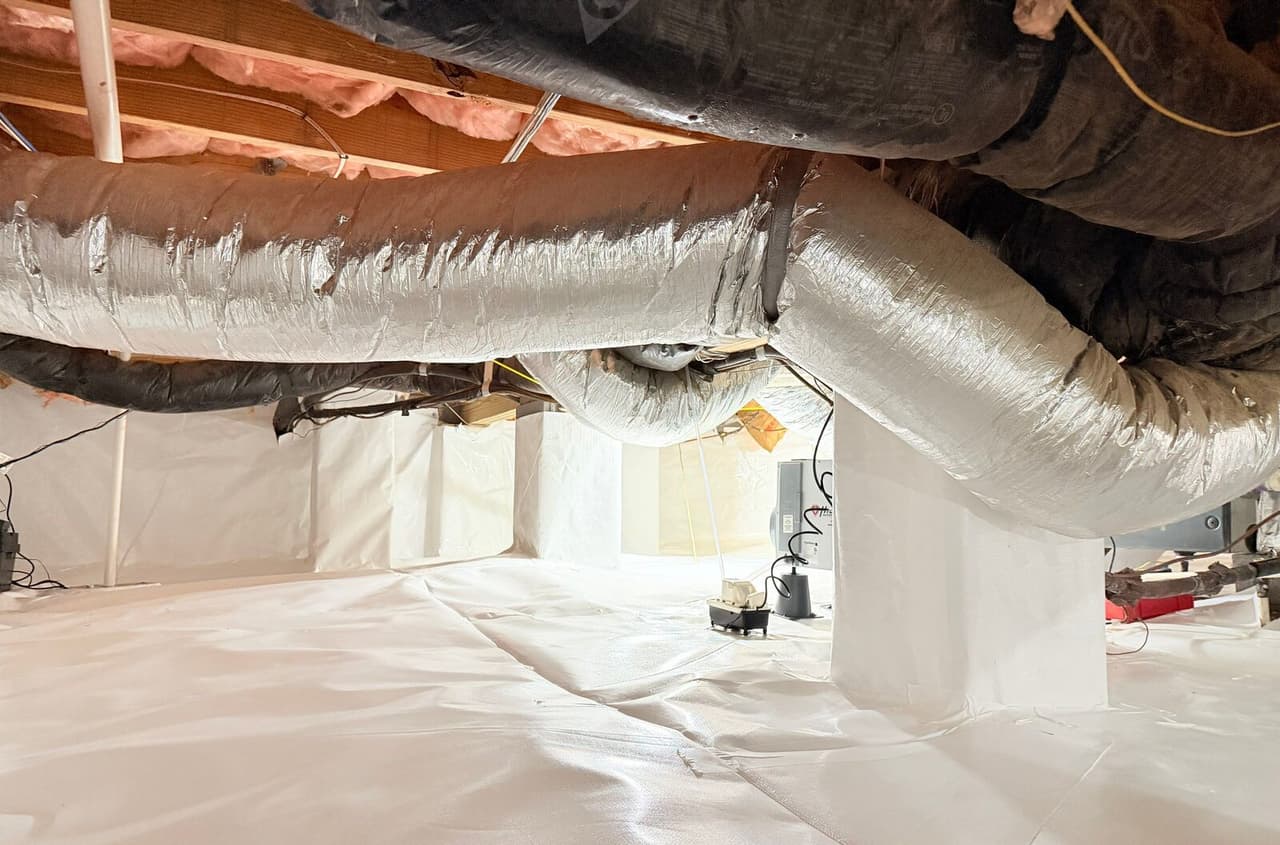 Lined crawl space with wrapped piers, insulated ducts and a pump unit on the white floor.