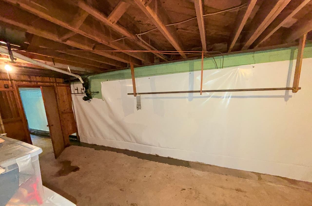 Long basement wall covered in white liner with a patched concrete strip at the base and exposed joists above.