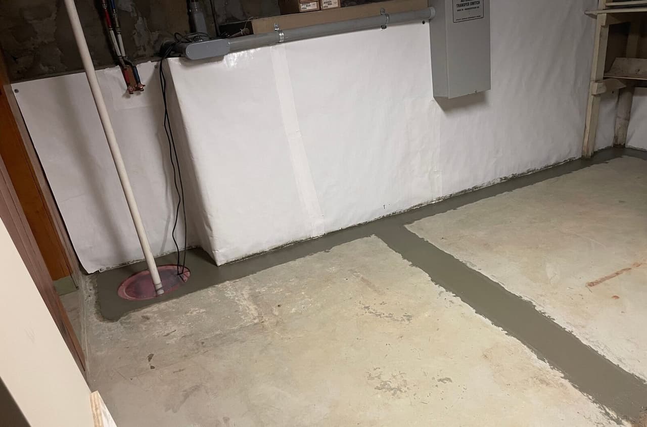 Basement corner with sump lid, discharge pipe, white wall liner and patched drain trench across the floor.