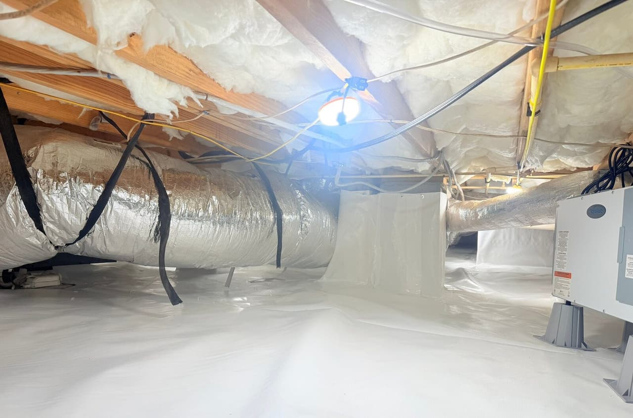 Wide view of a lined crawl space with a foil duct, work light, and dehumidifier at the right edge.
