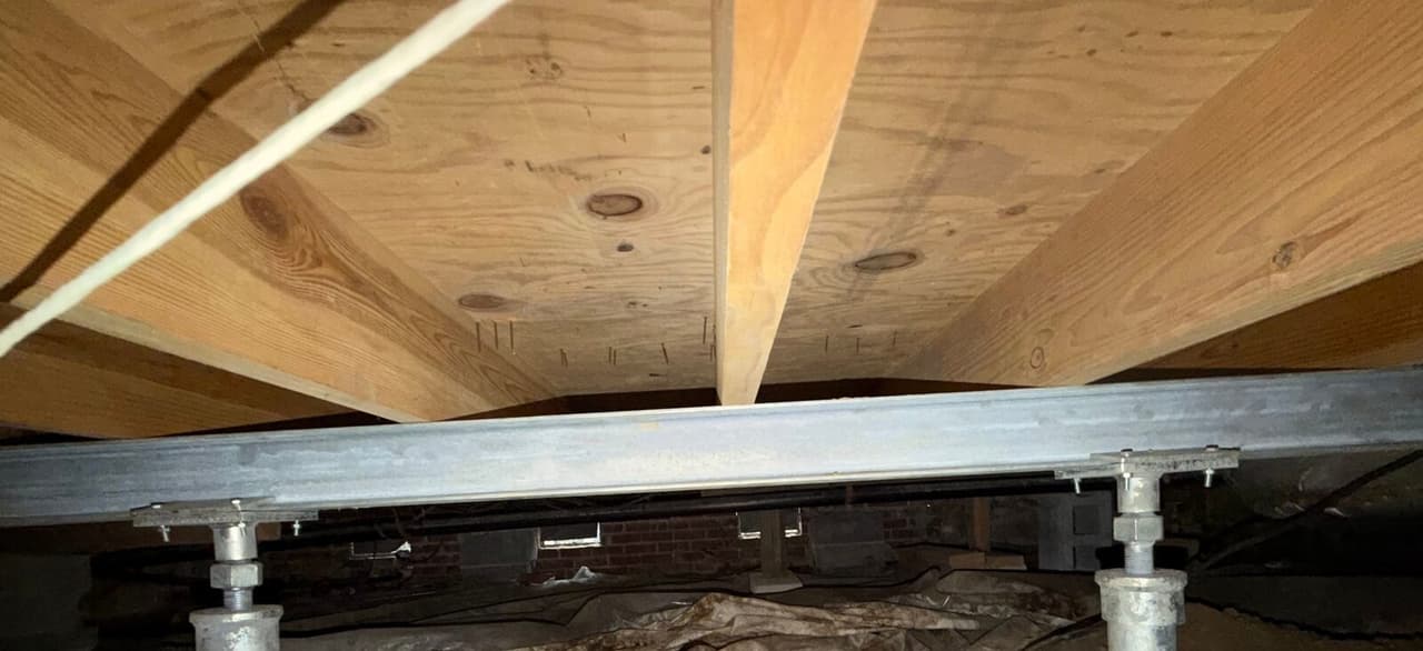 Steel beam supported by two adjustable steel posts under wood floor joists in a crawl space.