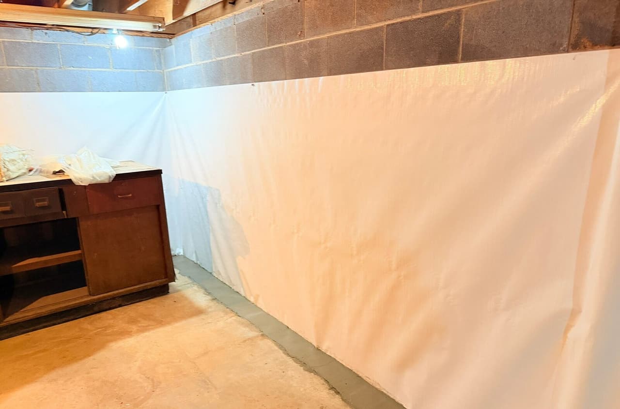 White waterproofing membrane on a block basement wall with a patched concrete strip along the floor edge.