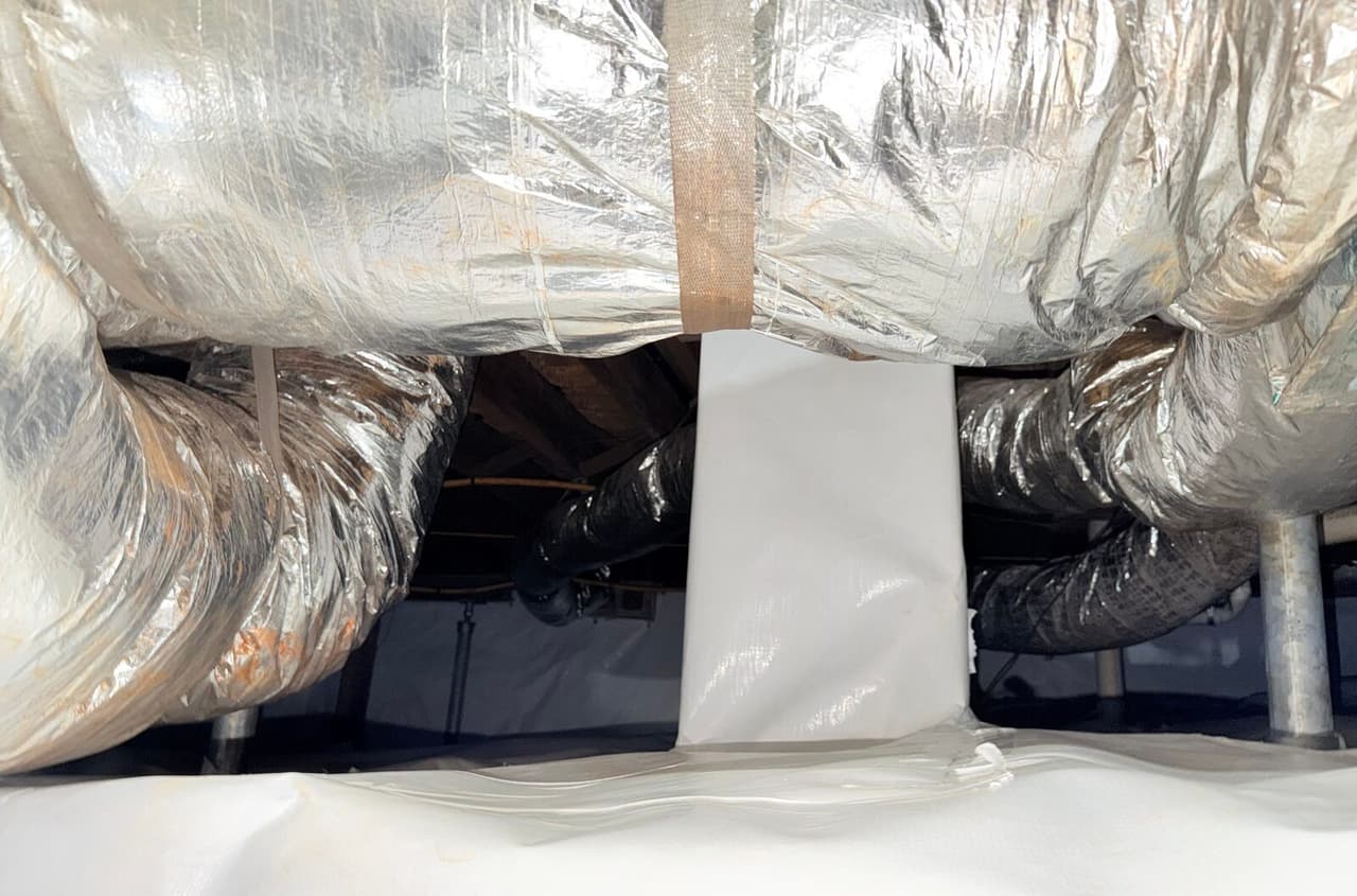 Crawl space with white liner covering the floor and a wrapped pier beneath foil wrapped ducts.